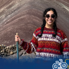 Smiling woman in colorful sweater holding hiking pole by banded red rock; small logo