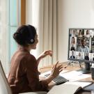 A person wearing headphones sits at a desk participating in a video conference call with multiple people displayed on a computer screen.