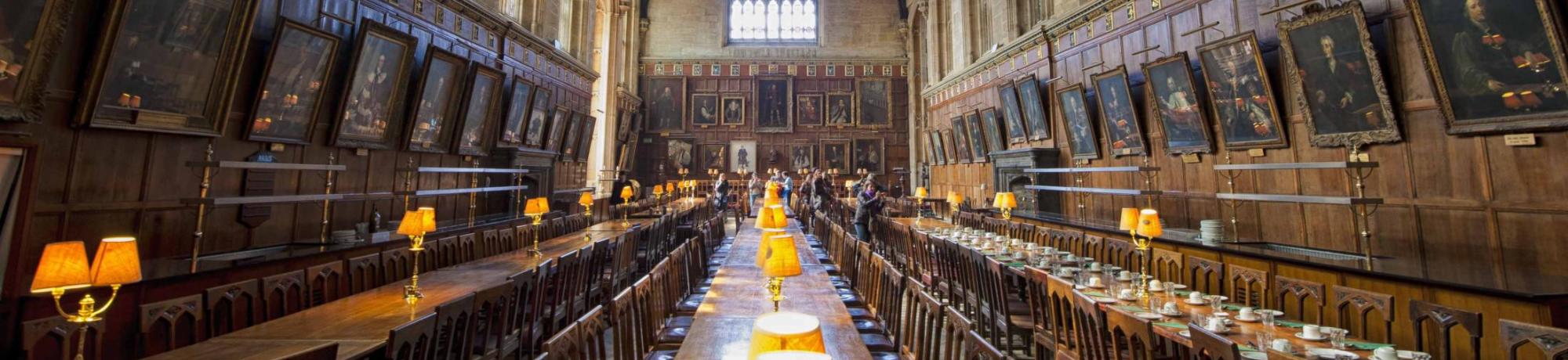 Long wooden dining tables in a grand, historic hall with high ceilings and large windows. Harry Potter-esque.