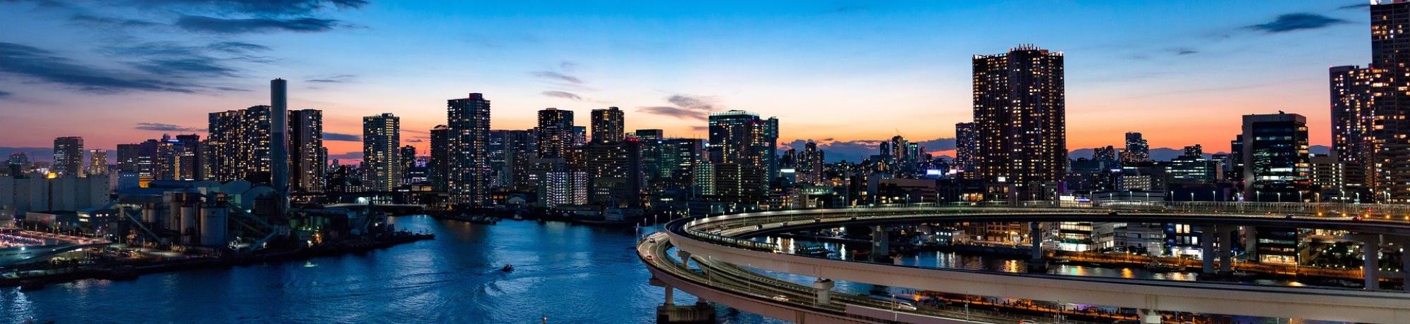 City skyline at sunset, with buildings reflecting on a river and a winding road.
